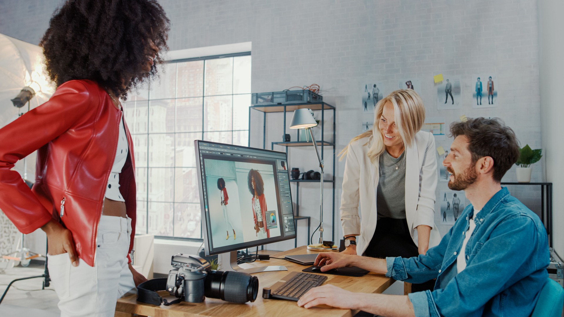 In Photo Studio Fashion Magazine Designer and Beautiful Black Cover Girl talk to a Professional Photographer who Uses Desktop Computer to Retouch Photographs in an Image Editing Software